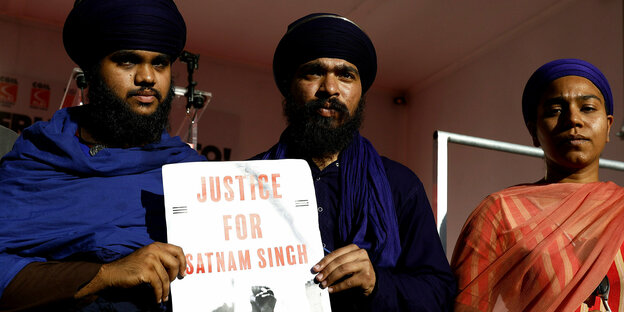 Zwei Männer und eine Frau in traditioneller indischer Kleidung stehen mit ersten Gesichtern auf einem Poedest, sie halten ein Plakat hoch: Justice for Satnam Singh