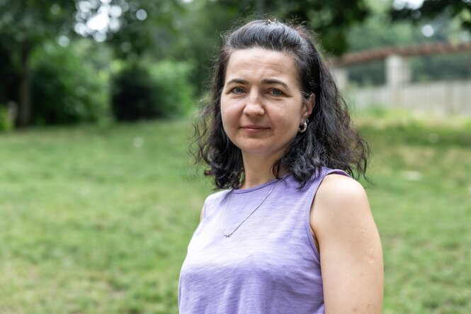 Eine Frau mit ärmellosem lila Shirt und langen scharzen Haaren schaut vor einer grünen Wiese