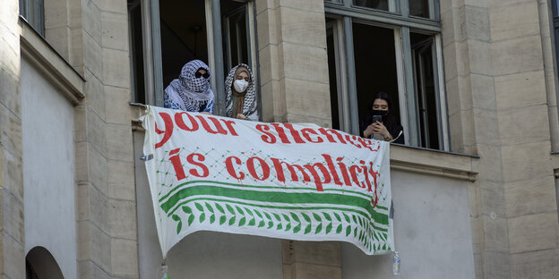 Besetzung der HU in Berlin: Studierende verdecken ihr Gesicht mit einer Kufiya, sie lehnen aus einem Fenster, an dem ein großes Transparent befestigt ist: Your Silence ist complicity (Dein Schweigen ist Komplizenschaft)