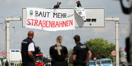 Drei Polizist*innen (von hinten fotografiert) stehen vor dem Hauptliefereingang von VW in Wolfsburg und beobachten, wie zwei Aktivist*innen Transparente vom Eingangstor herunterlassen. "Baut mehr Straßenbahnen" steht darauf