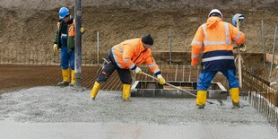 Drei Bauarbeiter mit Helm und Gummistiefel gießen Estrich