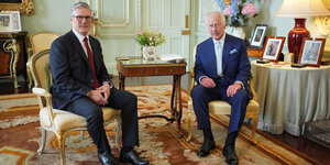 Keir Starmer und König Charles sitzen auf gepolsterten Stühlen, die golden lackiert sind, königliches Umfeld und schauen in die Kamera