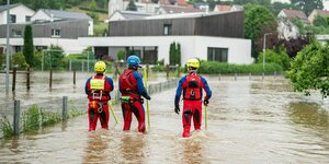 Drei Männer waten in Schutzkleidung durch eine überflutete Straße