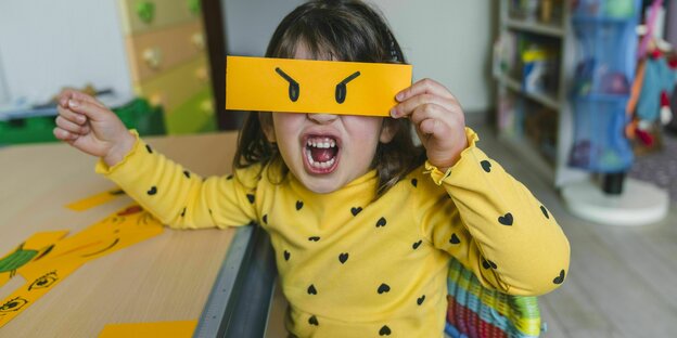 Kind macht ein wütendes Gesicht und hält dabei eine Maske mit wütenden Augen.