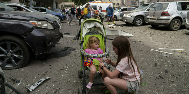 Ein größeres Kind kümmert sich um ein kleineres Kind im Kinderwagen, drumherum Trümmerteile.