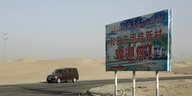 Das Ortsschild für ein uigurisches Dorf steht in einer wüstenartigen Landschaft der Region Xinjian in China, ein Auto kommt gerade aus der Richtung des Ortes