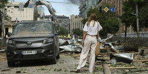 Eine Frau in heller Sommerkleidung steht auf einer Straße in Kyiv. Autos und Gebäude sind völlig zerstört