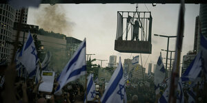 Eine Frau hängt in einem Käfig über einer demonstrierenden Menge. Darüber wehen Israelfahnen