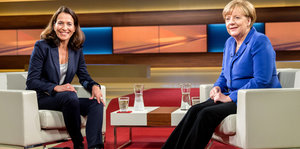 Eine dunkelhaarige Frau sitzt in einem Sessel, neben ihr auf einem Sessel Angela Merkel