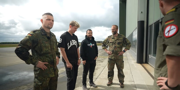 Drei Soldaten in Uniform und zwei junge Männer in schwarzen Hoodies stehen am Rand eines weitläufigen Geländes