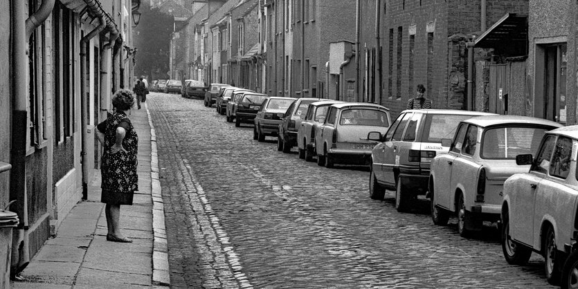 Gepflasterte Straße, eine Frau in Kittelschürze schaut die Straße hinunter, rechts parken Trabbis