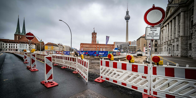 Ein Baustellenzaun an einer Straße, im Hintergrund der Berliner Fernsehturm