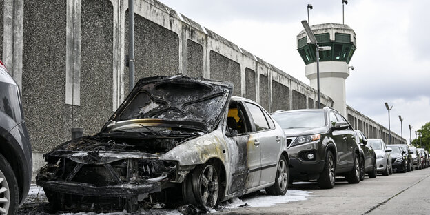 Ein ausgebranntes Auto steht vor einer hohen Mauer, im Hintergrund ein Wachturm