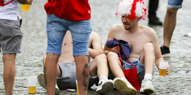 Zwei Männer mit nacktem Oberkörper sitzen auf dem Boden und trinken Bier, einer hat eine England-Flaggen-Perücke auf