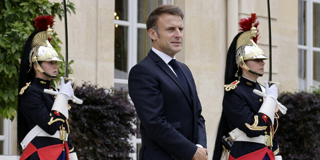 Emmanuel Macron steht vor dem Elysee Palast, zwei Wachmänner in altmodischen Uniformen stehen links und rechts von ihm