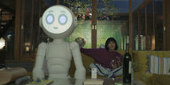 Ein Roboter sitzt auf dem Sofa, hinter ihm eine Frau in einem Wohnzimmer