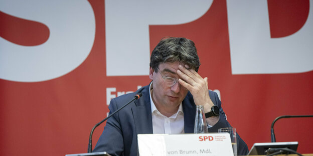 Florian von Brunn sitzt auf dem Podiumvor einem großen SPD Logo und stützt seinen Kopf in die Hände