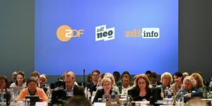 Der ZDF Fernsehrat: Menchen sitzen an Plätzen und schauen auf ihre Labtops und im Hintergrund eine blaue Wand mit den Logos von ZDF, zdf neo und zdf info