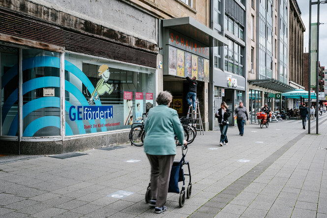 Passanten in einer Straße, im Vordergrund eine Frau mit Rollator