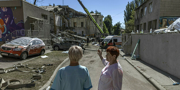 Zwei Frauen in Arbeitskitteln stehen auf einer Straße, im Hintergrund sieht amn ein völlig zerstörtes Haus