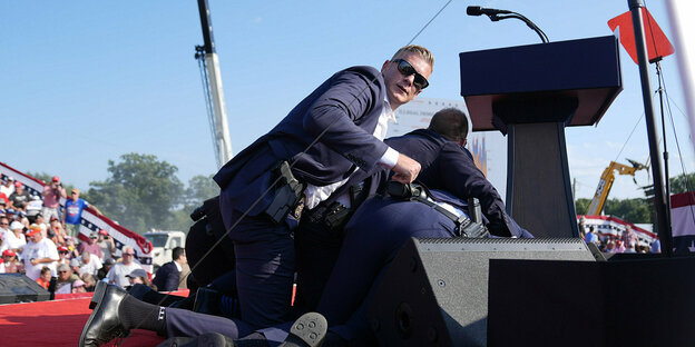 Ein Sicherheitsmann des US-Geheimdienstes kniet auf einer Bühne. Er trägt eine Sonnenbrille und blickt direkt in die Kamera