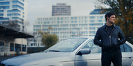 Trojan (Mišel Matičević) steht vor einem silbernen BMW auf einer anonymen Gewerbefläche.