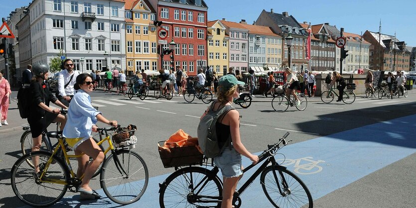 Viele Menschen fahren mit Fahrrädern über eine Kreuzung an einer Brücke, einige Radfahrer:innen warten auf einem farbig markierten Radweg. Die kleine Brücke führt auf eine Straße, die von bunten Häusern gesäumt ist.