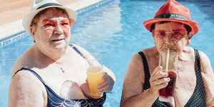 Zwei weiße Frauen im Badeanzug schauen in die Kamera. Im Hintergrund erkennt man unscharf einen Pool. Beide tragen Sonnenhut, Sonnenbrille. Beide halte in einer Hand ein Getränk. Die Frau zur Rechten trinkt gerade durch einen Strohhalm.