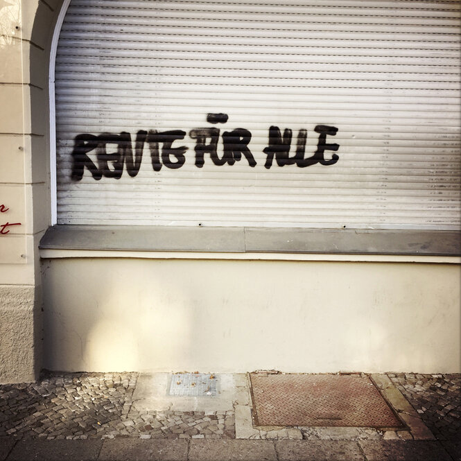 Auf weißen Rolladen, neben einer Hauswand, steht "Rente für alle", in schwarz, gesprüht.