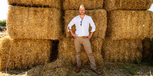 Der Landwirt Benjamin Meise steht in weißem Hemd und beigfarbener Hose vor einem Stapel Strohballen