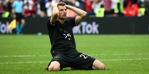 Thomas Müller kniet auf dem Rasen im Stadion, die Arme hinter dem Kopf verschränkt und schaut etwas entgeistert