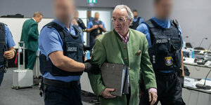 Prinz Reuß zwischen zwei Polizisten im Gerichtssaal, er trägt ein grünes Jackett sowie einen Aktenordner