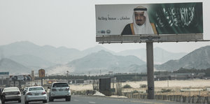 drei Autos auf einer Straße, darüber auf einem Werbeplakat ein Mann neben arabischen Schriftzeichen
