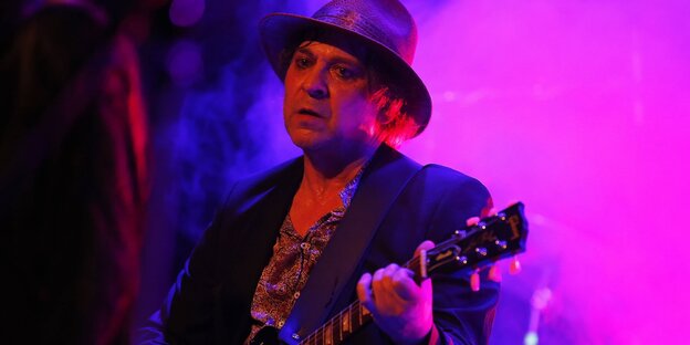 R. P. S. Lanrue steht mit der Gitarre auf einer Bühne vor lila Nebel.
