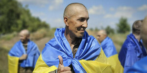 Ein ukrainischer Kriegsgefangener mit ein Daumen-hoch-Zeichen, und einer Ukraine Flagge über den Schultern nach einem Gefangenenaustausch. Im Hintergrund stehen weitere entlassene Männer mit umgelegten Fahnen