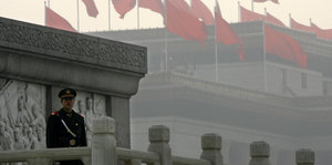 Ein Offizier steht auf einem Balkon eines Nachtbargebäudes des Nationalen Volkskongress Chinas. Im Hintergrund ist das Gebäude des Volkskongresses zu sehen, mit etlichen chinesischen Flaggen, die vom Smog vernebelt sind.