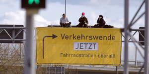 Klimaaktivist*innen auf einer sogenannten Schilderbrücke an einer Autobahn; das ursprüngliche Autbahnschild ist mit einem Transparent in ähnlicher Optik überklebt, darauf steht "Verkehrswende jetzt! Fahrradstraßen überall"