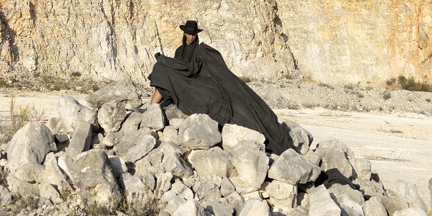 Eine Frau sitzt mit langem schwarzen Umhang und schwarzem Zylinder auf einem Felsen