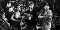 Schwarz-weiß-Screenshot aus dem Film Road To Bali aus dem Jahr 1952: Ein Mensch mit Hose, Hemd und Schiebermütze steht am Strand vor Palmen. Er hält eine kleine Schatzkiste unter dem Arm, zwei andere Person rechts und links von ihm gucken erschrocken in d