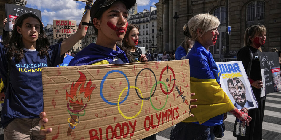 Auf einer Demonstration zu Ehren der getöteten ukrainischen Sportler hält einer der Demonstranten ein Pappschild, auf dem blutbespritzte olympische Ringe zu sehen sind. Darunter die Worte "Bloody Olympics".