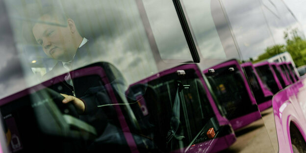 Mehrere pinke Busse stehen nebeneinander auf einem Parkplatz. Sie sind durch die Windschutzscheibe eines weiteres Busses fotografiert, in der sich ein Busfahrer spiegelt. Er trägt eine DB-Uniform und hat die Hand am Lenkrad.