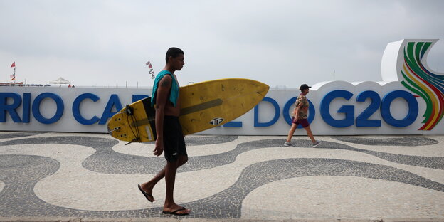 Ein Mann mit Surfbrett geht in Rio de Janeiro vor dem offiziellen G20 Logo