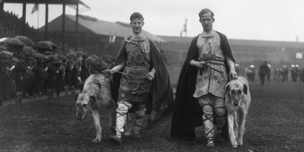 Eine schwarz-weiß Aufnahme zweier junger Männer in traditionell irischen Gewändern, die jeweils einen Hund an der Leine führen
