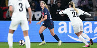 Eine blonde, junge Fußballspielerin in blauen Trikot spielt den Ball gegen zwei Spielerinnen in Weiss