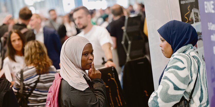 ZweiFrauen mit Kopftuch stehen auf einer Messe vor einer Wand