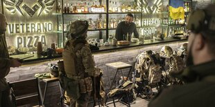 Ein Mann steht hinter einem Tresen, hinter ihm ein Regal mit Flaschen und einer ukrainischen Fahne, vor dem Tresen Menschen in Flecktarn: Pizzeria Veterano in Kyjiw