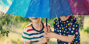 Zwei Kinder stehen lachend, unter einem bunten Regenschirm, im Regen.
