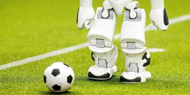 Die Beine und der angeschnittene Oberkörpers eines kleinen Roboters, der auf einem gründen Fußballfeld steht und vor ihm liegt ein Fussball