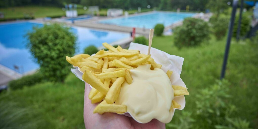 In der Hand eine Schale mit Pommes und viel Majo - im Hintergrund siend die Becken eines Freibades zu sehen