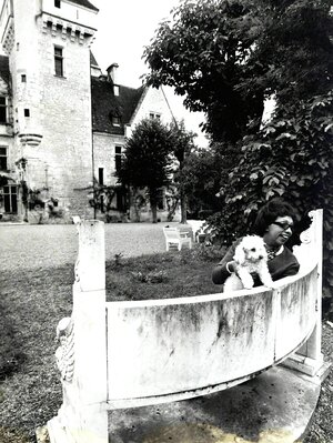 Josephine Baker sitzt mit einem hellen Pudel auf einer steinernen Bank vor dem Schloss. Sie trägt eine dunkle Hornbrille und lacht